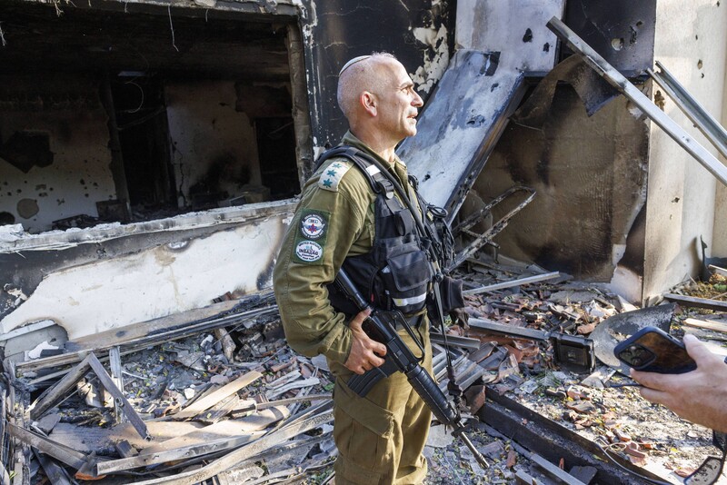 Man in military uniform stands in front of badly burned and damaged house