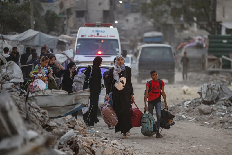 Women and children carry belongings amidst rubble. Ambulances are behind them. 