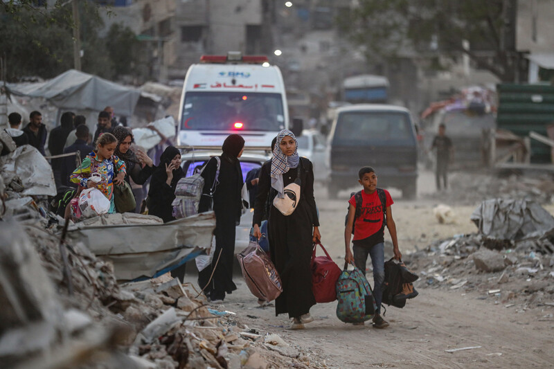 Women and children carry belongings amidst rubble. Ambulances are behind them.