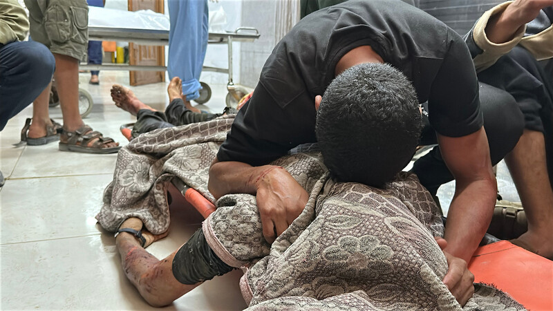 A man cradles the body of a loved one on the floor of a hospital. The body is wrapped in a blanket.
