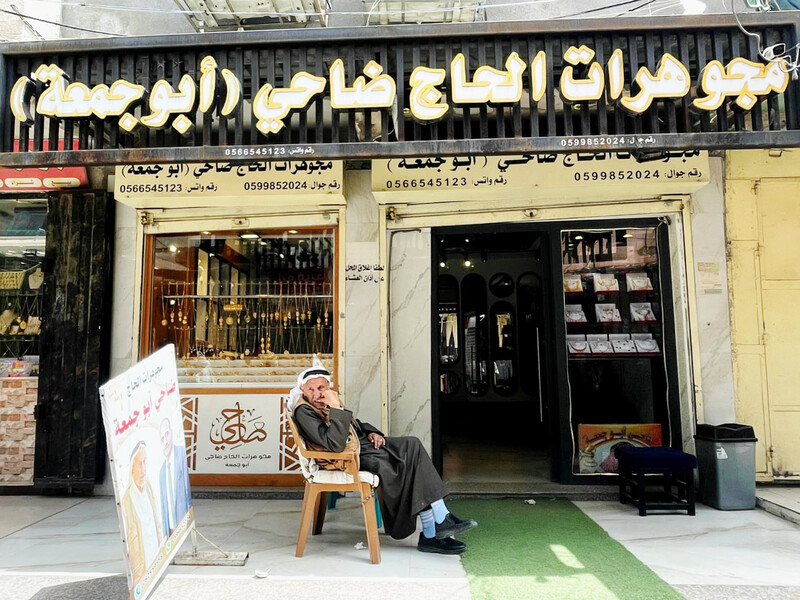 An older man sits in a plastic chair in front of a jewelry shop