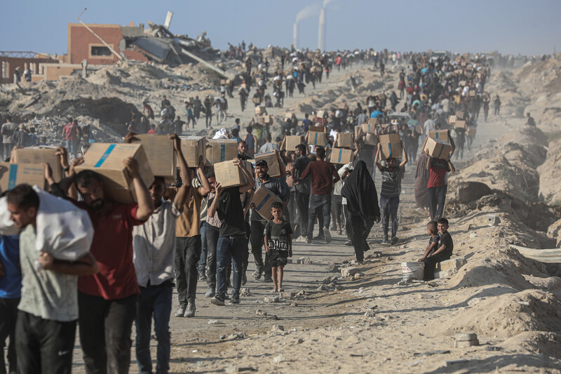 Scores of men walk on dusty road while carrying boxes on their heads and shoulders