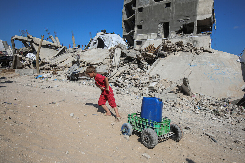 A child, dressed in red, pulls a can of water on a small cart up a hill, surrounded by rubble of destroyed buildings.