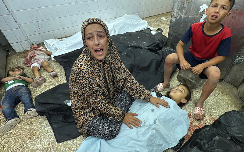 A woman wearing prayer clothes mourns over the body of a small child under a blue medical blanket. Two small children lie motionless behind her as an older boy sits next to her.