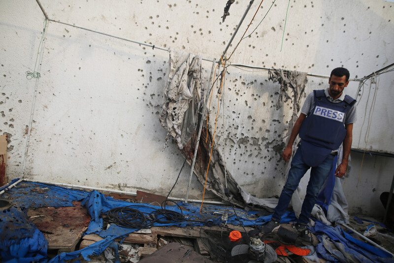 A man in a press vest looks down at the damage after an airstrike. There are shrapnel holes in the wall beside him.