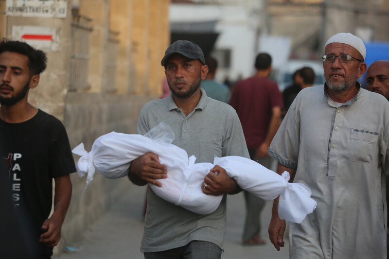 A man holds the body of a child, wrapped in a white burial shroud. Two men walk alongside him