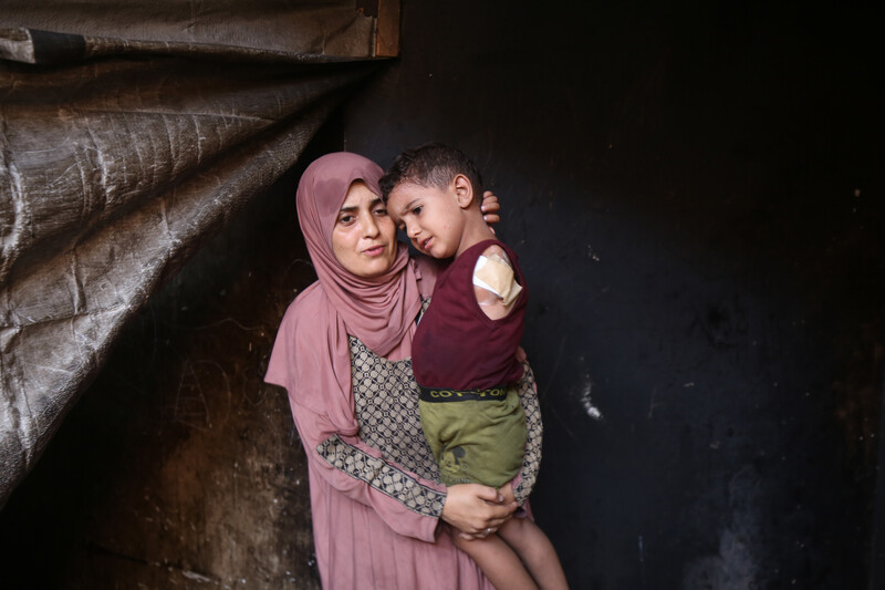 A woman wearing pink prayer clothes holds a toddler, dressed in a red shirt and green shorts. The toddler is missing his left arm.
