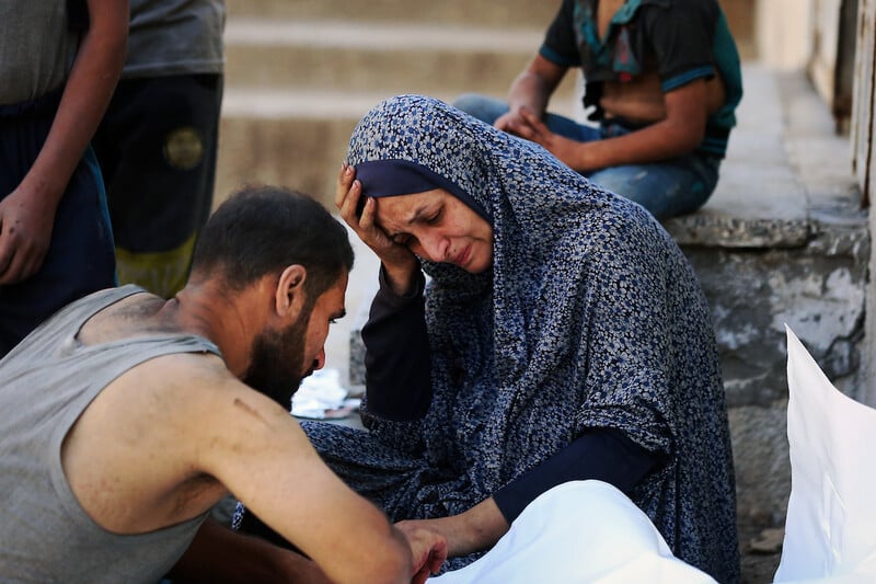 A woman in a blue headscarf holds her head in her hand. A man faces her. They are both looking at a body wrapped in a white shroud.