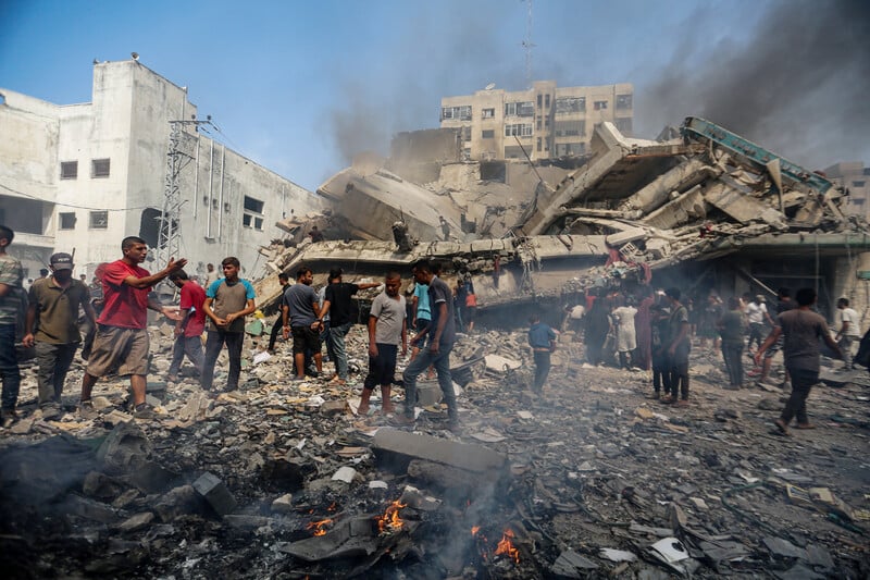 People stand on top of the rubble of a destroyed building, with black smoke rising around them. Several small fires are visible in front of them.