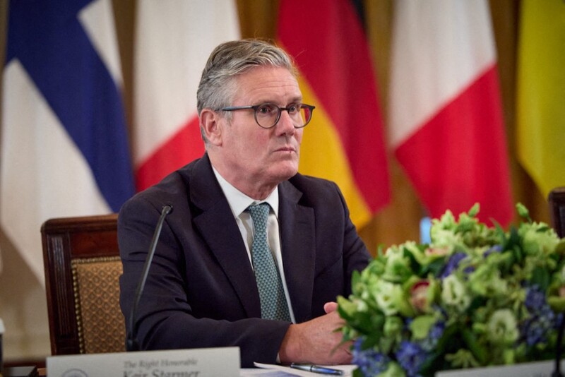 Keir Starmer sits at a table with flowers in front of him and flags behind him