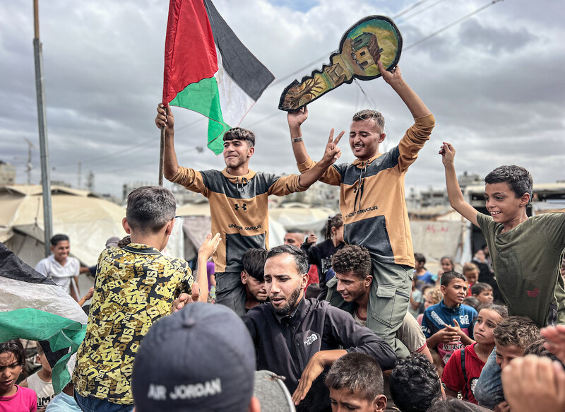 A group of young Palestinian men and children stand in a group. Carried on shoulders, two men hold a Palestinian flag and a symbolic key. Everyone is smiling.