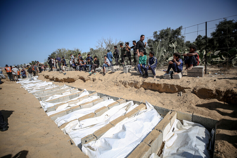 People sit next to a row of graves, filled with bodies in white shrouds