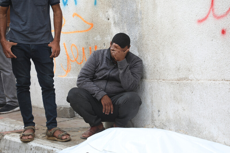 A man in a grey jacket and dark pants crouches against a wall, next to a body wrapped in a white shroud. His hand is covering his face.