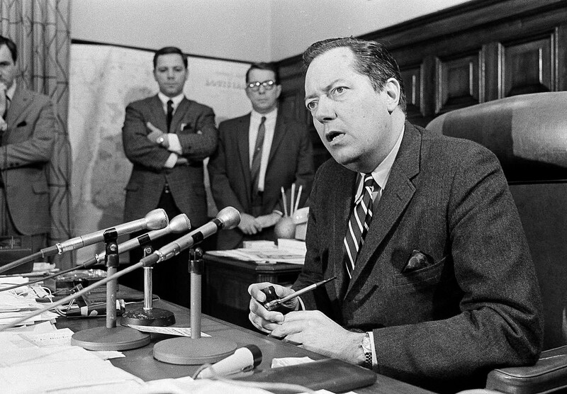 A man at a desk in a suit speaking into microphones