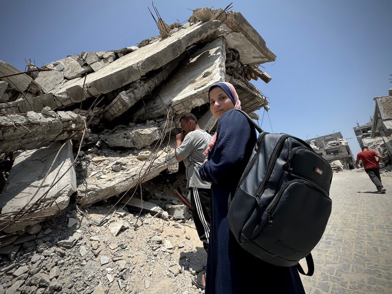 A teenage girl in a headscarf carrying a backpack and posing to the camera while her father stands behind her and looking at the rubble of a completely bombed house.