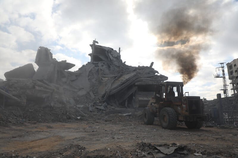 A bulldozer clears rubble from a destroyed house with smoke still billowing in the background