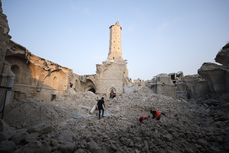 A few number of people inspecting heaps of rubble with a partially destroyed minaret standing.