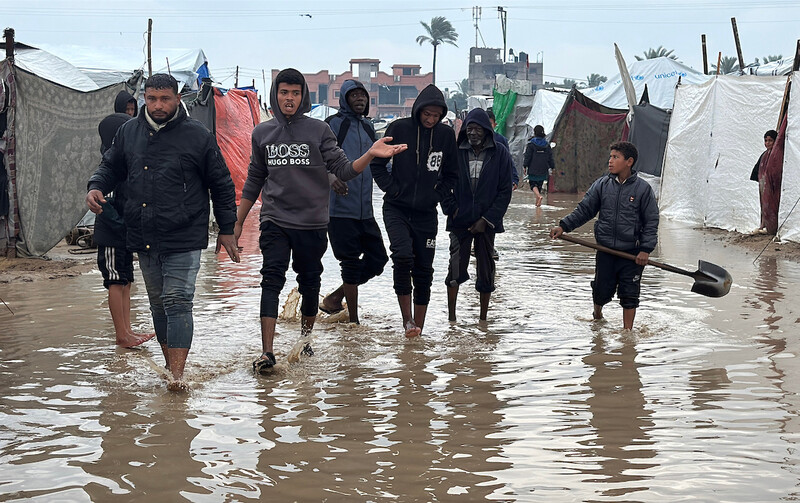 A group of youth walk through a flooded street between tents. One of them is carrying a shovel.