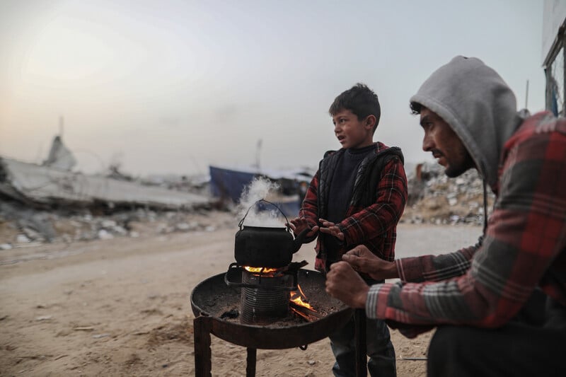 A man and a boy warm their hands over a small tray with a fire and a teapot over it