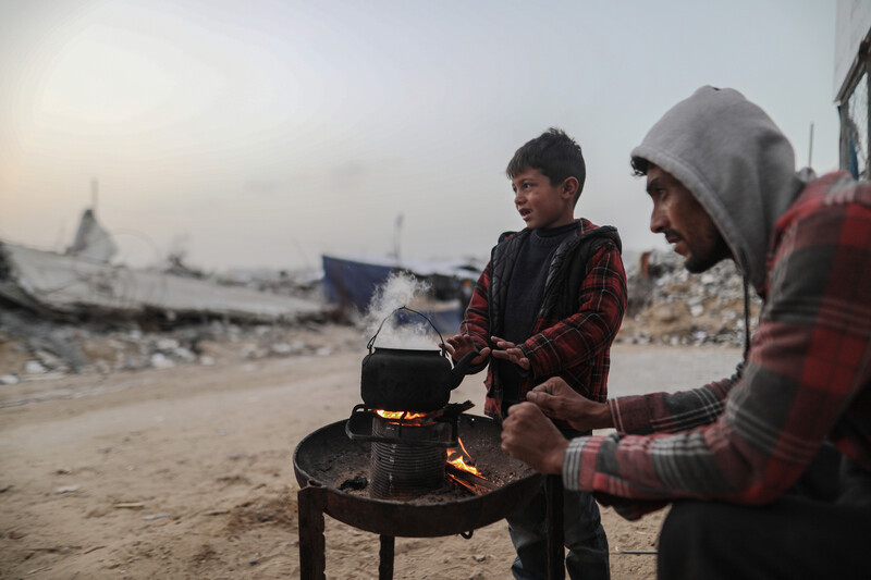 A man and a boy warm their hands over a small tray with a fire and a teapot over it