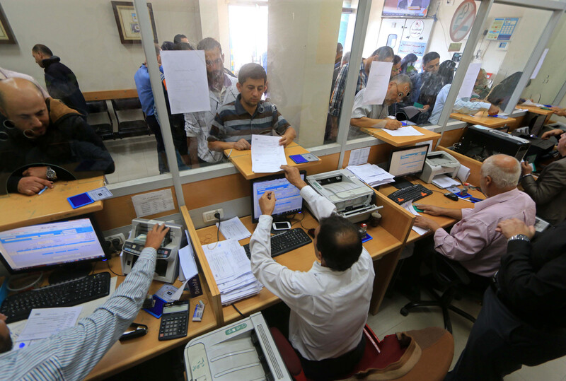 Workers sit at a partitioned counter with computers while people queue in front of them