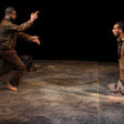 A standing man gestures to a kneeling man on a dark, spare stage