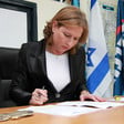 Tzipi Livni sits at desk looking at paper with an Israeli flag hanging behind her
