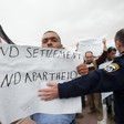Israeli police officer places arm across Palestinian protester carrying sign reading End Settlements and Apartheid
