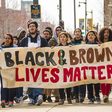 Racially diverse group of young people carry banner reading Black and Brown Lives Matter 