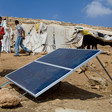 A boy stands next to a solar panel installed on barren ground with makeshift shelters in the background