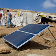 A boy stands next to a solar panel installed on barren ground with makeshift shelters in the background