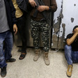 Photo shows mourning man sitting on the ground with his head in his hands as three men, one of them carrying a rifle, standing nearby are shown from waist down