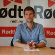 Young man speaks while sitting at desk with a Rødt party banner behind him