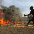 A youth pulls on a barbed wire fence that is partially set on fire as a crowd looks on