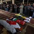 Men stand with their arms folded across their torsos in front of six shrouded bodies