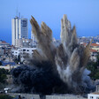 Smoke rises over buildings against Gaza skyline. 