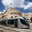 A tram passes through a street with sandstone buildings behind it