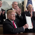 Four standing men applaud another man sat at a desk holding up a piece of paper.