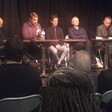 Five men, one with a microphone, sitting behind a table