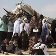 Women and children stand and sit in front of rubble of destroyed structure