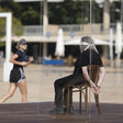 Blindfolded woman cuffed to a chair sits in a glass box 