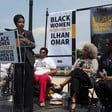 Woman speaks at podium with women and banners behind her