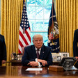 Man sits behind a desk, two men stand behind him 
