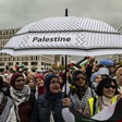 A group of women stand beneath a black-and-white umbrella emblazoned with the word 'Palestine'. 