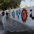 Men use hoses to spray sidewalk along wall with mural depicting a face in a coronavirus sphere and surgical masks 