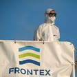 Man wearing mask and other protective equipment stands behind Frontex banner
