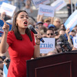 Woman speaks at podium in front of large crowd carrying signs 