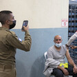 A uniformed soldier holds up a phone while a seated man receives a shot from a health care worker