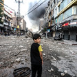 A boy stands in debris-strewn street with smoke on the horizon
