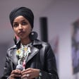 Ilhan Omar, wearing leather jacket, stands while holding microphone